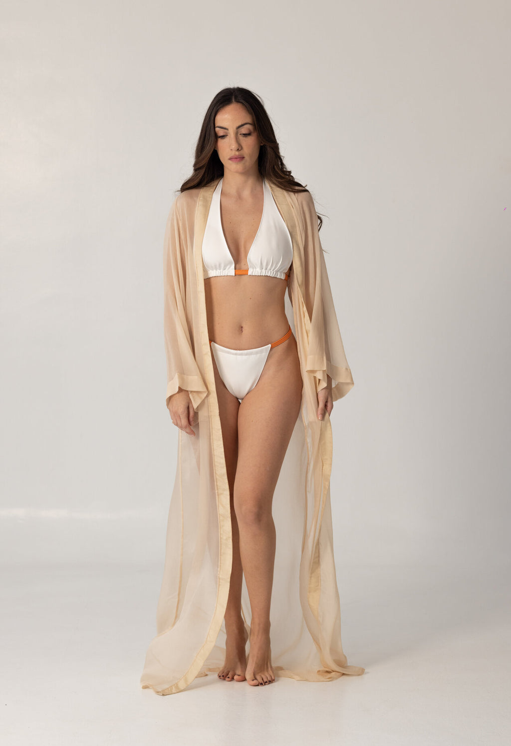 Woman wearing a white and orange bikini with a sheer beige cover-up standing against a plain background showcasing the ISAURA luxury swimwear style.