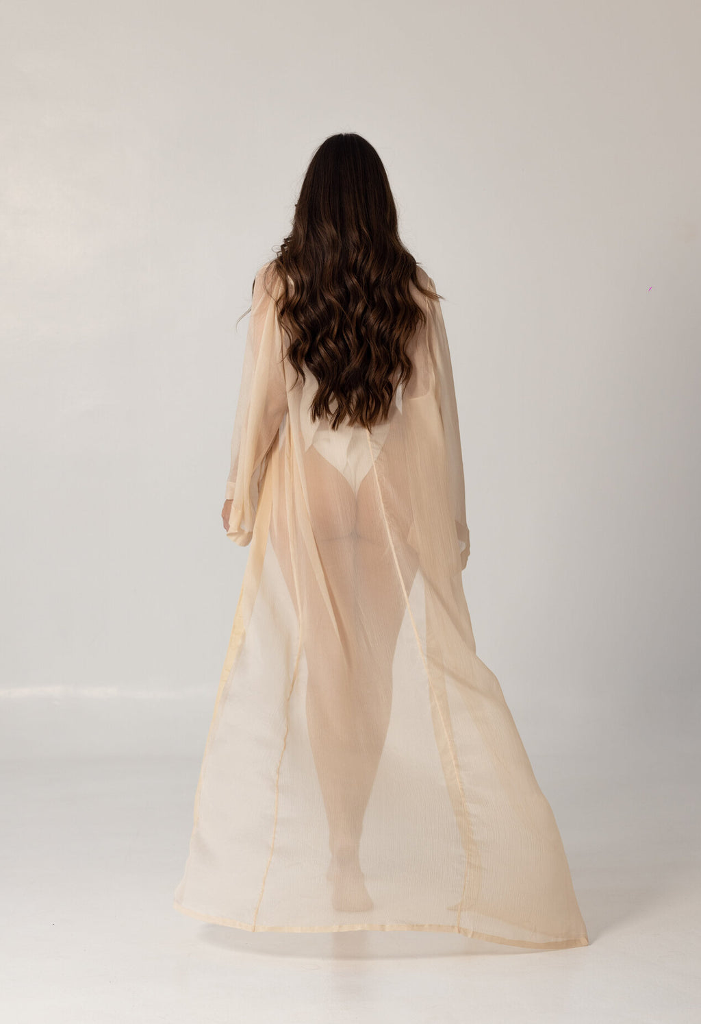 Model wearing a sheer, flowing beige beach cover-up from behind, showcasing the elegant and modest design of the ISAURA luxury swimwear.