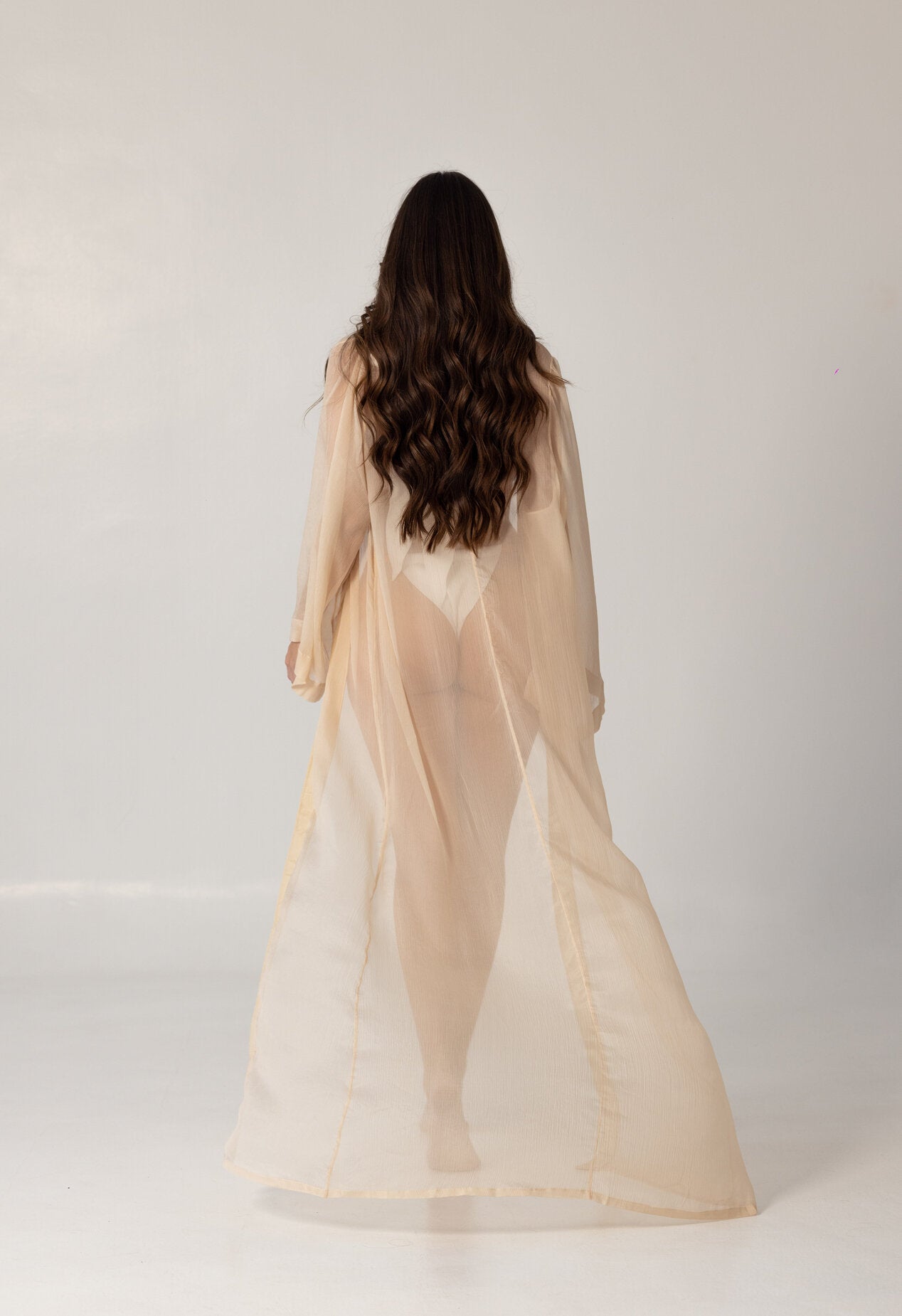 Model wearing a sheer, flowing beige beach cover-up from behind, showcasing the elegant and modest design of the ISAURA luxury swimwear.
