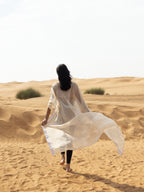 Woman walking barefoot in a sandy desert wearing a flowing sheer beach cover-up named ISAURA, styled as modest luxury resort wear with a light, airy fabric.