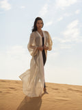Woman wearing a flowing sheer beige cover-up called ISAURA over a black outfit, standing barefoot on sand dunes under a bright sky.