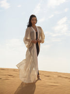Woman wearing a flowing sheer beige cover-up called ISAURA over a black outfit, standing barefoot on sand dunes under a bright sky.