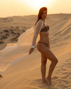 Woman wearing a stylish JANNAH bikini standing on sand dunes at sunset with a flowing sheer cover-up, showcasing luxury swimwear in a desert setting.
